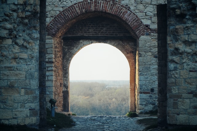 Through an old stone archway, a bright valley filled with trees is visible.