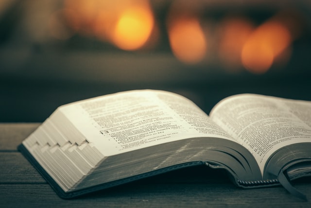 An open bible rests upon a wooden table with a campfire out of focus in the background.