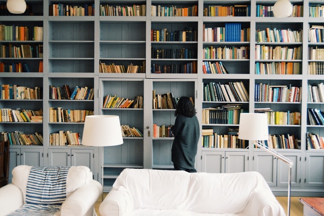 A person stands next to a couch and looks up toward a wall of books.