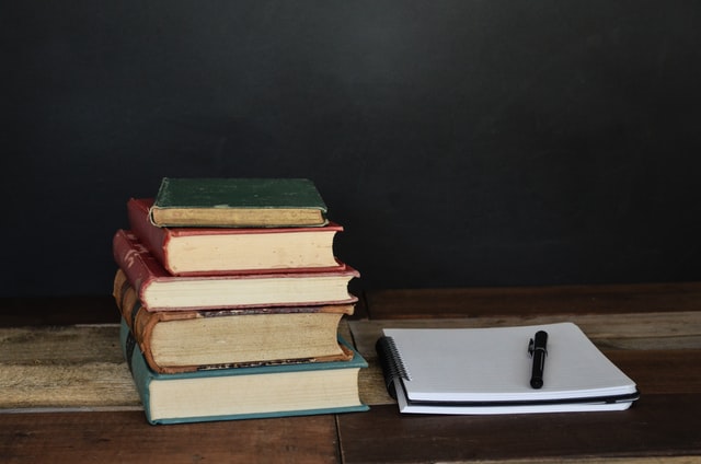 An open notebook, a pen, and a stack of old hardcover books rest upon a wooden table.