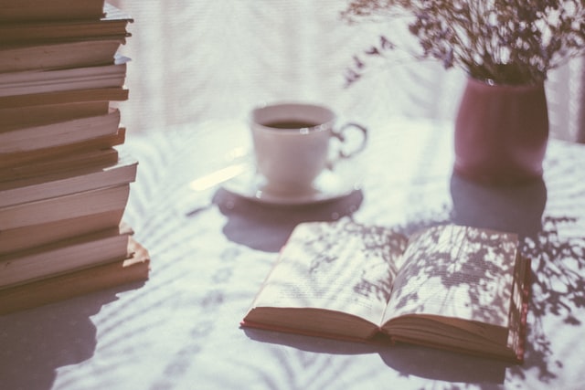 An open book rests upon a table next to a stack of books and a cup of tea. Sunlight shines upon the table.