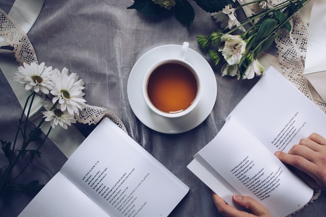 A person reads a book on a table with tea and flowers.