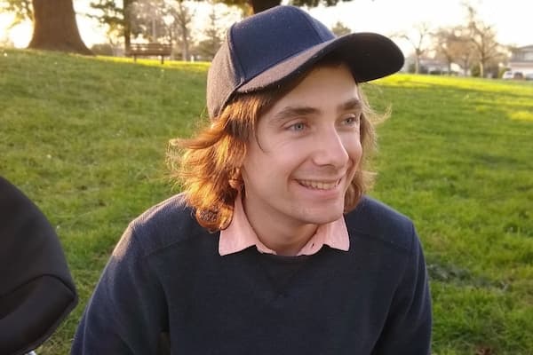 Cam Coulter smiles at a park on a sunny day. They wear a navy ballcap and sweater and a salmon-colored button-up.