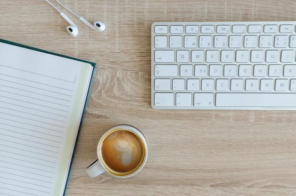 A keyboard, a notebook, headphones, and a cup of coffee rest upon a wooden desk.